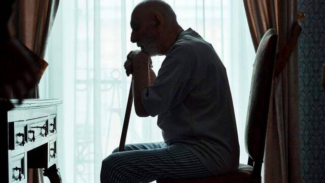 Un anciano apoyado en un bastón, sentado, a contraluz de una ventana.