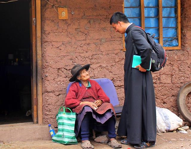 Un sacerdote con sotana conversa con una mujer anciana a la que visita.