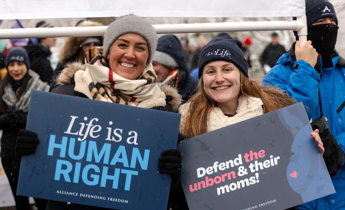 Dos mujeres sonrientes y abrigadas con carteles provida en la Marcha por la Vida 2024 de Washington