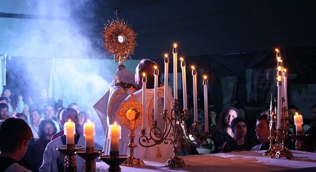 Sacerdote eleva la Custodia en Adoración eucarística con velas, foco e incienso