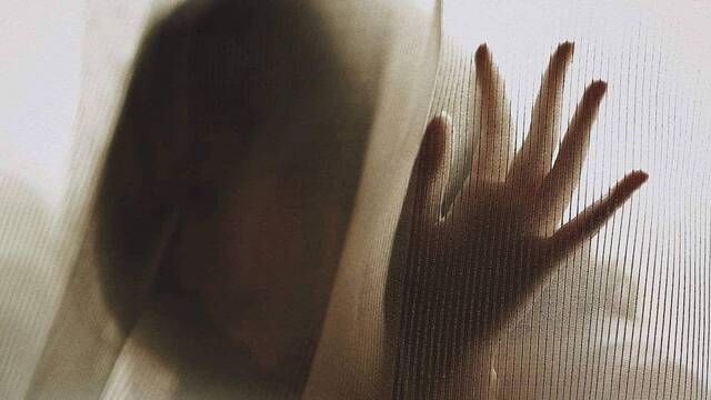 Mujer triste tras una ventana en la que apoya la mano.