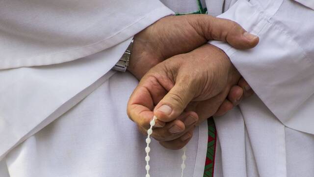 Manos de un sacerdote revestido rezando el rosario.