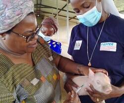Sor Christabel -con mascarilla- sostiene un cerdito que vacuna su superior, Sor Rosalia Sakayombo, en su granja-escuela premiada