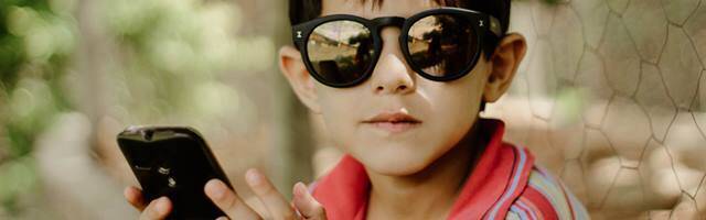 Niño pequeño con gafas de sol y un teléfono móvil en las manos mira a cámara.