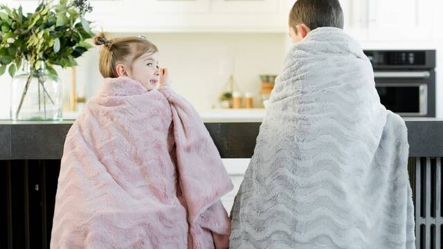 Dos niños de espaldas, en la cocina abrigados con mantas.