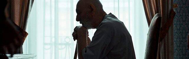 Un anciano apoyado en un bastón, sentado, a contraluz de una ventana.