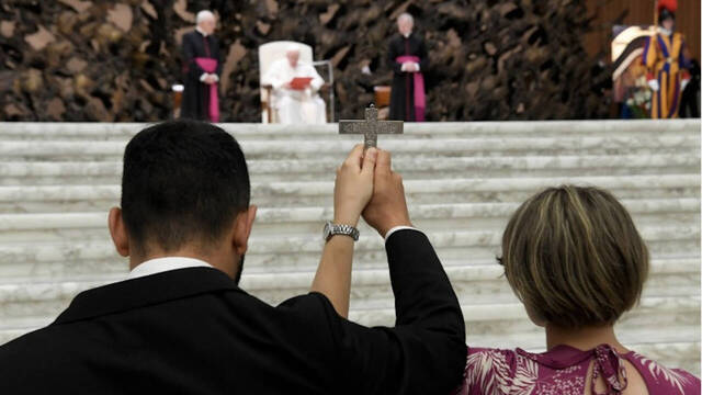 Un hombre y una mujer sostienen juntos una cruz con el Papa Francisco al fondo.