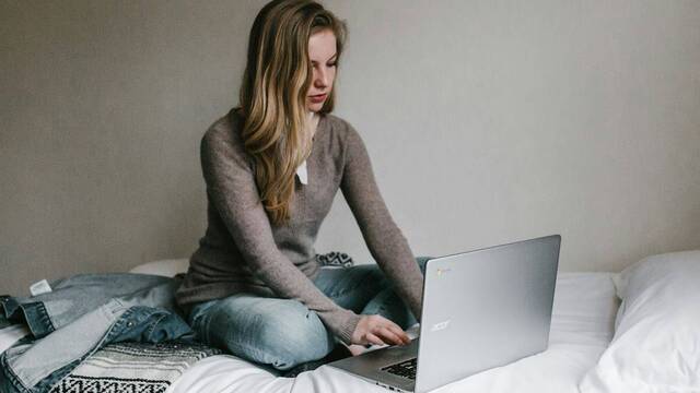 Una joven escribe sobre un portátil sentada en su cama.