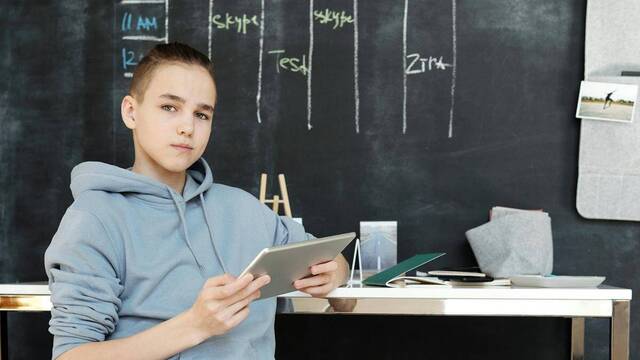 Un alumno adolescente mira a cámara con una tablet en las manos, en el aula.