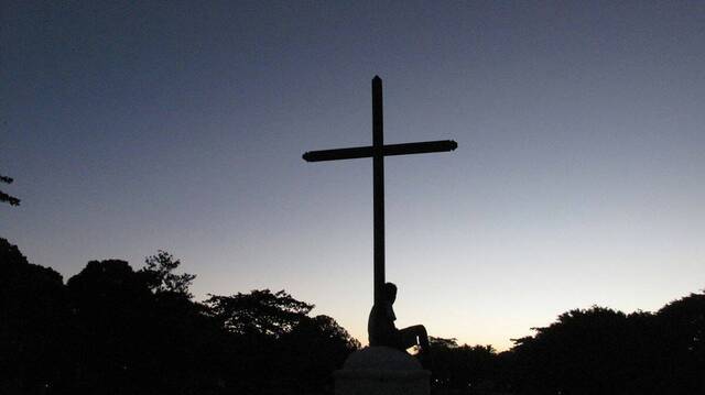 En contraluz, una mujer sentada a los pies de una cruz en la montaña.